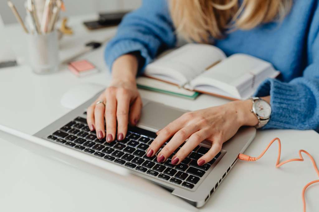 Frau mit blauem Pullover, deren Hände auf einer Laptop-Tastatur zum Tippen ansetzen. Vor ihr liegen aufgeschlagene Notizbücher.