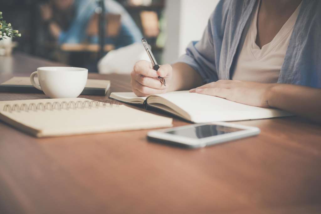 Schreibtisch mit dunklem Holz, Kaffeetasse, Smartphone, Notizbücher, Person mit weißem Shirt und hellblauem Cardigan, die in Notizbuch schreibt,