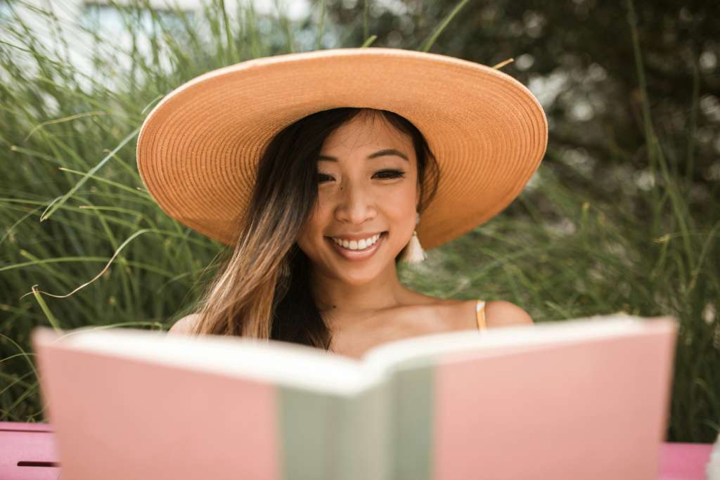Lächelnde Frau mit Sonnenhut liest in rosafarbenem Buch