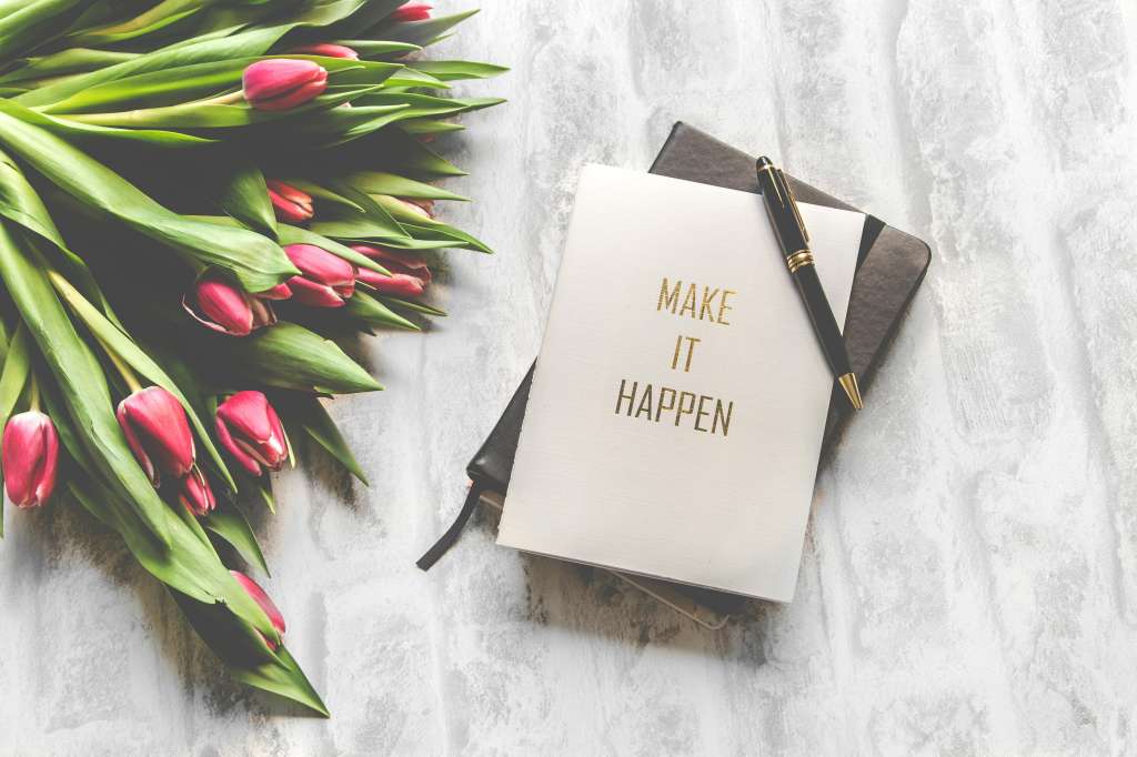 Notizbuch mit der Aufschrift ‚Make it Happen‘, umgeben von frischen Tulpen und einem Stift – sinnbildlich für das Schreiben und Veröffentlichen eines Buches als Selfpublisherin.